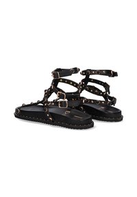 Sandalias negras con tiras, que cuentan con múltiples hebillas ajustables, tachuelas doradas y negras, suela plana y una textura en la suela para mayor agarre.