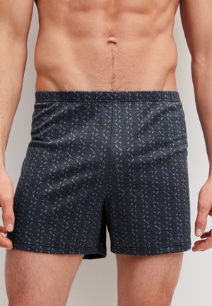 Männlicher Torso, der schwarze Boxershorts mit kleinem blauem geometrischem Muster trägt, vor einem einfarbigen Hintergrund.