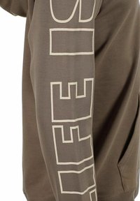 Felpa marrone con una grande grafica testuale color crema lungo la manica. Tessuto morbido con una vestibilità comoda e una tasca frontale.