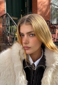 Zatvorenim planom osobe s dugom, ravnom plavom kosom, koja nosi kaput s kragnom od krzna boje krem na tamnoj jakni i bijeloj košulji s kravatom.