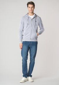 Felpa grigia con cappuccio e zip, dotata di lacci, abbinata a jeans in denim blu e sneakers bianche. Il tessuto è morbido e ha una vestibilità casual.