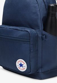 Sac à dos bleu marine en tissu résistant, avec une poche avant zippée, un patch rond avec le logo Converse, et une poche latérale pour une bouteille d'eau.