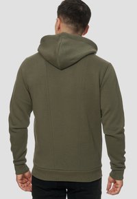 Sudadera con capucha de color verde oliva que presenta una textura suave, puños y dobladillo acanalados, y un diseño aerodinámico con detalles en las costuras traseras.