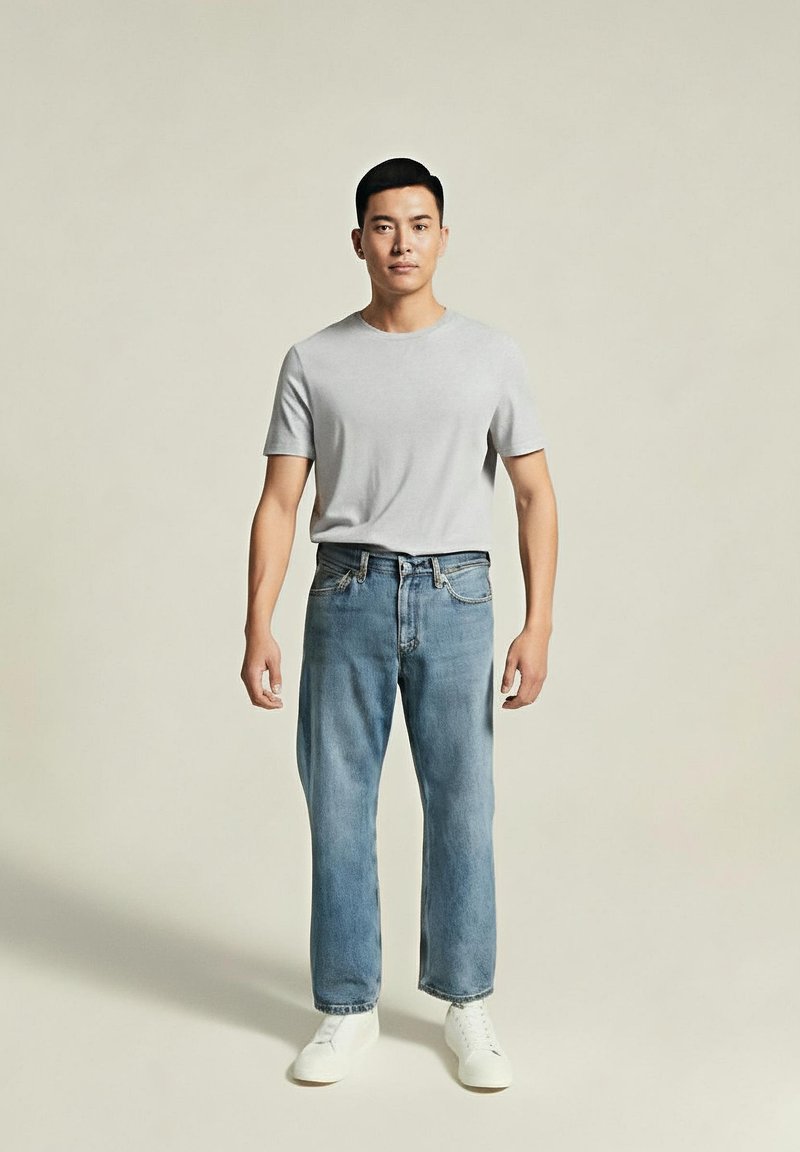 Hombre de pie contra un fondo liso, vistiendo una camiseta gris claro, jeans azules y zapatillas blancas, mirando hacia adelante con una postura relajada.