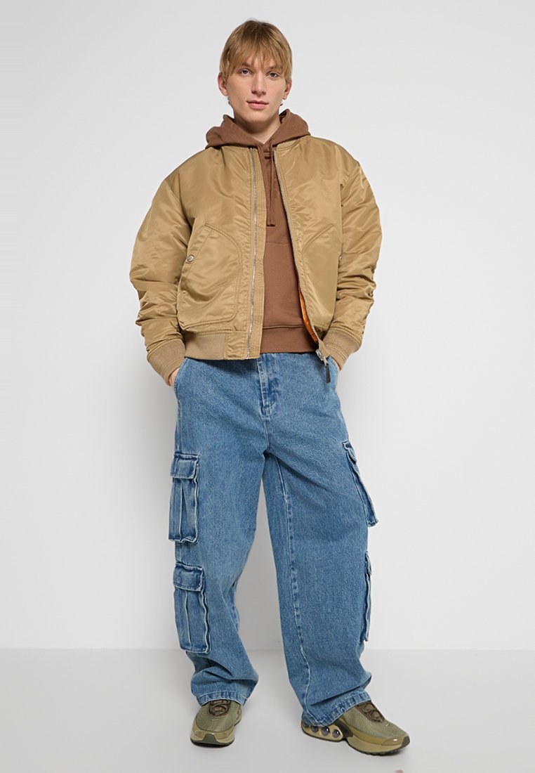 Khaaki kurtka bombowa z prążkowanymi mankietami na brązowym hoodie, w połączeniu z luźnymi niebieskimi spodniami cargo z wieloma kieszeniami. Oliwkowe trampki.