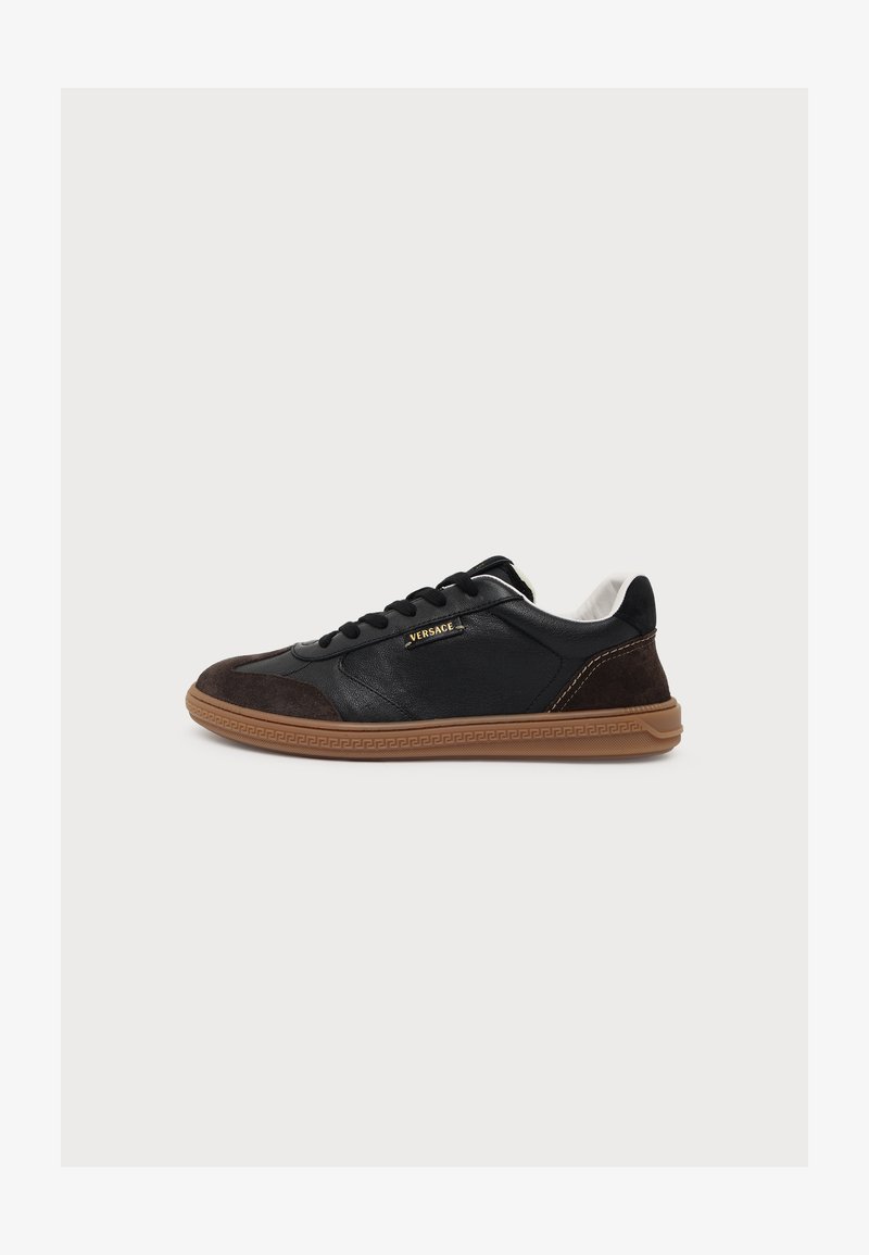 Basket noire Versace avec des accents en daim marron, logo doré sur le côté et semelle en caoutchouc marron, présentée de profil sur un fond uni.
