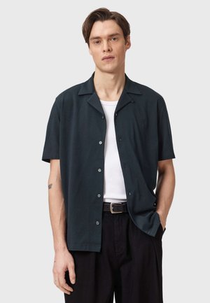 Junger Mann mit kurzen braunen Haaren trägt ein schwarzes, kurzärmeliges Hemd über einem weißen T-Shirt, schwarze Hose mit Gürtel, steht mit einer Hand in der Tasche.