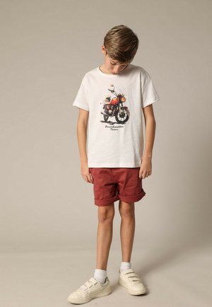 T-shirt blanche avec un motif de moto, associée à un short rouge, des chaussettes blanches et des chaussures blanches. Tenue simple et décontractée.
