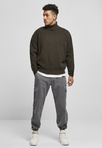 Brun strikket turtleneck sweater, grå cargo bukser med lommer og hvide high-top sneakers. Løs pasform, struktureret stof og lag-på-lag look.