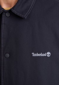 Black button-up jacket made of durable fabric, featuring a classic collar and white embroidered "Timberland" logo with a tree icon.