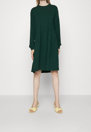 Personne portant une robe longue à manches longues de couleur vert foncé, arrivant au genou, et des sandales à talons ouverts jaunes, se tenant devant un fond blanc uni.