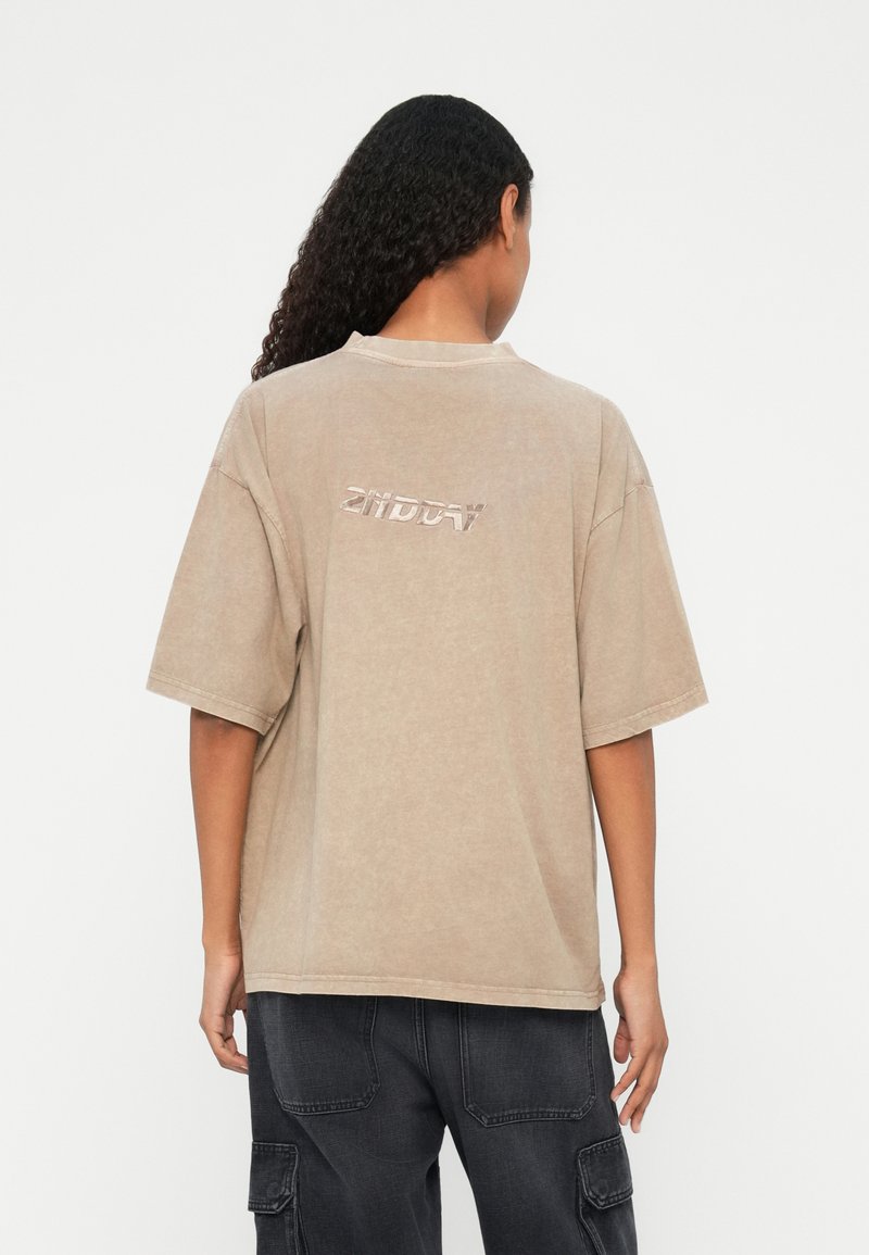 Beige oversized t-shirt med rund halsudskæring og korte ærmer. Har et skinnende, præget logo på ryggen. Glat tekstur, afslappet design.