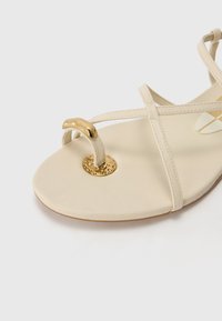 Sandalo in pelle beige con delicate cinghie incrociate, un accento decorativo in tono oro e una suola rotonda e testurizzata.