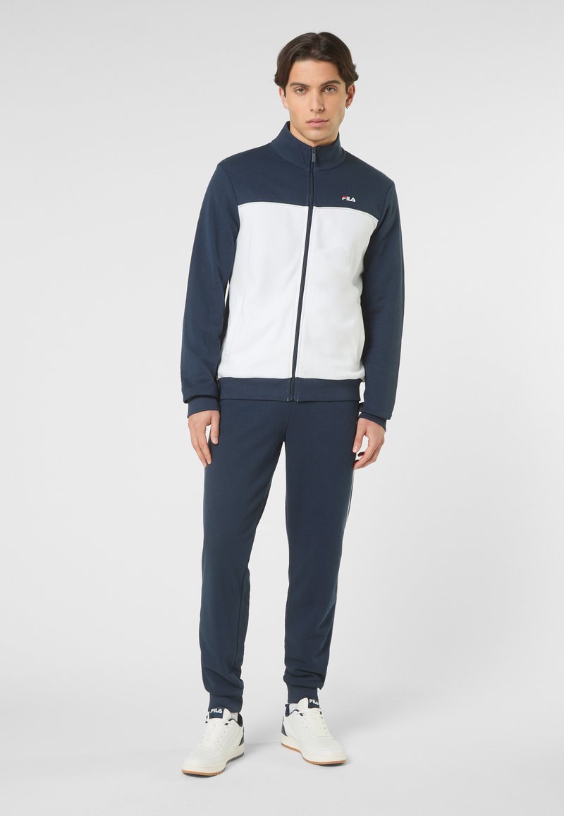 Jeune homme debout portant un survêtement zippé bleu marine et blanc avec un pantalon bleu marine et des baskets blanches FILA sur un fond uni.