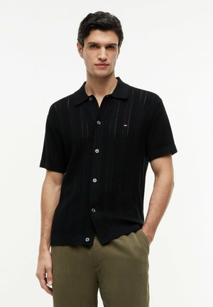 Uomo che indossa una maglia nera a maniche corte con bottoni e pantaloni verde oliva, in piedi con una mano in tasca su sfondo bianco.