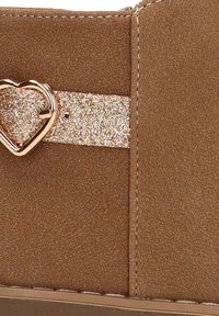 Botín de ante marrón con una correa dorada brillante que presenta un acento metálico en forma de corazón. Detalles de costura visibles en la parte superior.