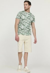 Chemise verte à manches courtes avec un motif de feuilles, associée à un short beige clair. Baskets couleur ivoire avec des accents clairs.