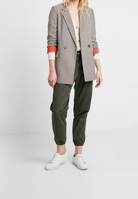 Geruite blazer met oranje manchetten, lichte button-up, olijfgroene tapered broek en witte sneakers.