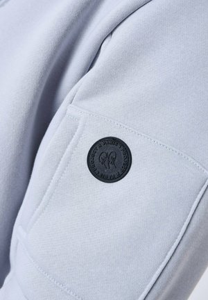 La tela del sudadera gris claro presenta un bolsillo cosido con un parche de logo negro redondo que muestra detalles de diseño y texto.
