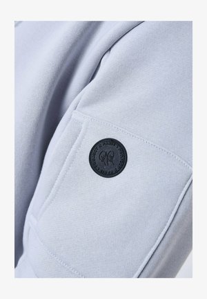 Il tessuto del felpa grigio chiaro presenta una tasca cucita con un'etichetta rotonda nera che mostra dettagli del design e testo.