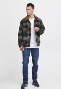 Flanellen jas in groen, zwart en beige ruitpatroon; lange mouwen, knoopsluiting aan de voorkant, één borstzak. Witte t-shirt en blauwe jeans eronder gedragen.