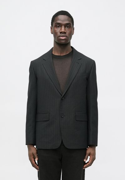 Schwarzer Blazer mit Nadelstreifen und figurbetontem Schnitt, zwei vorderen Taschen und notched Revers, getragen über einem dunklen, strukturierten Oberteil.