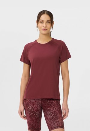 Femme portant un t-shirt uni bordeaux à manches courtes et un short bordeaux à motifs jusqu'aux genoux, debout devant un fond gris clair.