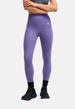 Kobieta nosząca fioletowe legginsy z wysokim stanem i boczną kieszenią, czarne skarpetki oraz czarne sportowe buty, stojąca z założonymi rękami.