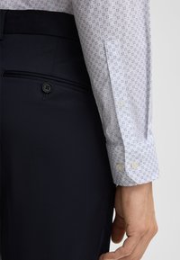 Witte blouse met een klein marineblauw geometrisch patroon, gecombineerd met marineblauwe broek. De broek heeft een knoop en plooidetail; de blouse heeft knoopmanchetten.