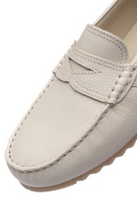 Bežová kožená mokasína s detailním stehováním, průřezovým páskem přes horní část a texturovaným okrajem podrážky v detailním záběru.