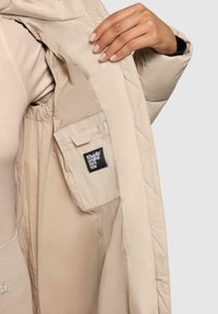 Beige gewatteerde jas binnenkant met een elastische manchet, voorzien van een zijzak en een merklabel dat aan de binnenkant is gestikt. Gladde textuur.