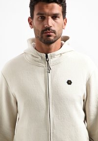 Beige textuur zip-up hoodie met een hoge kraag, verstelbare trekkoordkap en een klein zwart hexagonaal logo op de borst.