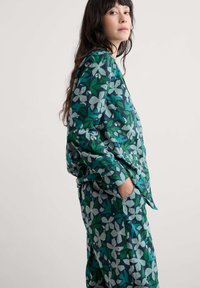 Lillelille mustriga koduriietus, millel on suured rohelised ja sinised lehed koos valgete õitega, valmistatud pehmest kangast, lõdva istuvusega ja küljetaskutega.