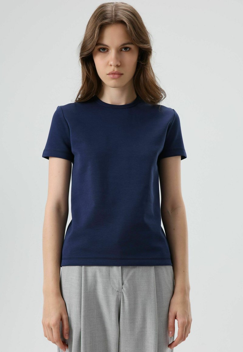 Marineblauw T-shirt met korte mouwen gemaakt van gladde stof; ronde hals, aansluitend model en subtiele rekbaarheid; gecombineerd met lichtgrijze broek.