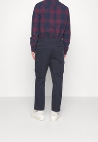 Pantaloni cargo navy con gamba dritta, dotati di tasche laterali e una finitura leggermente testurizzata, abbinati a una camicia a quadri in rosso scuro e navy.