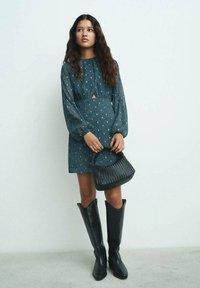 Teal-mönstrad klänning med nyckelhålsutskärning, långa ärmar och en avslappnad passform; i kombination med svarta knähöga stövlar och en vävd handväska.