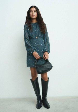 Abito teal con motivo a fantasia, con scollatura a chiave, maniche lunghe e vestibilità rilassata; abbinato a stivali neri al ginocchio e una borsa intrecciata.