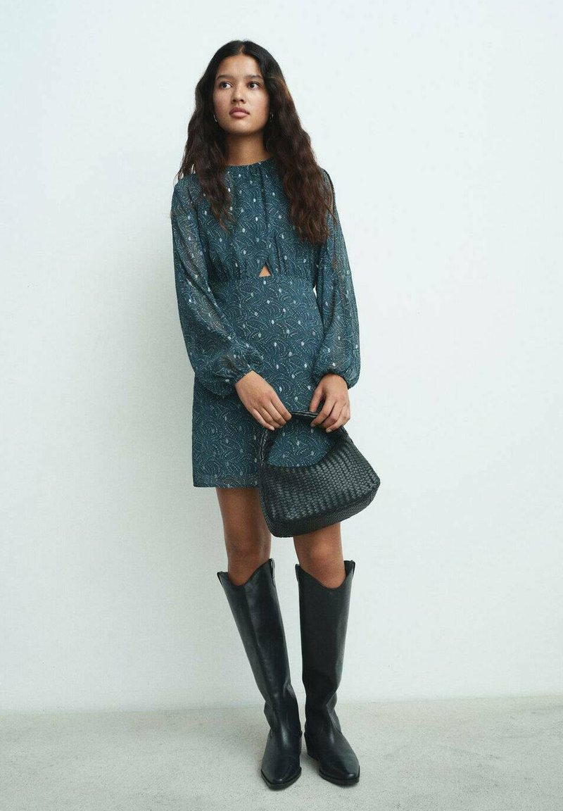 Teal-mönstrad klänning med nyckelhålsutskärning, långa ärmar och en avslappnad passform; i kombination med svarta knähöga stövlar och en vävd handväska.