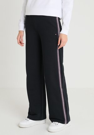 Person iført sorte bukser med vide ben med hvide og pink sidelister, hvide sneakers og en hvid langærmet top.