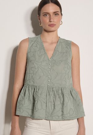 Mujer con blusa sin mangas verde salvia texturizada con botones y bajo con volantes, y pendientes de aro dorados, de pie frente a un fondo claro.