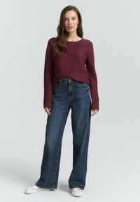 Weinroter gerippter Strickpullover mit langen Ärmeln, kombiniert mit weit geschnittenen dunkelblauen Denim-Jeans und weißen Sneakern.