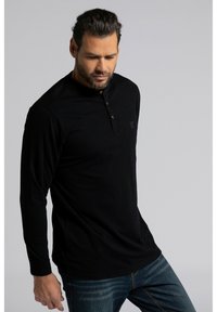 Schwarzes Langarm-Henley-Shirt mit drei Holzknöpfen und einem dezenten Logo auf der Brust, kombiniert mit blauen Denim-Jeans.