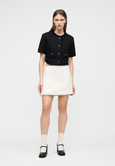 Γυναίκα με μαύρο κοντομάνικο κοντό σακάκι με διακοσμητικά κουμπιά, άσπρη υφασμάτινη μίνι φούστα, μπεζ κάλτσες και μαύρες γόβες Mary Jane.