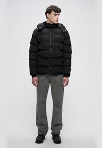 Geacă matlasată neagră cu fermoar frontal, glugă și quilting orizontal. Asortată cu pantaloni cargo gri și cizme negre, pe un fundal simplu.