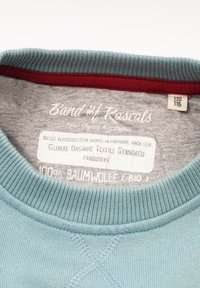 Vaaleansininen puuvillaneule, jossa resorireunainen pääntie; harmaa vuori ja punainen yksityiskohta. Etiketissä ilmoitetaan orgaanisen materiaalin noudattaminen.