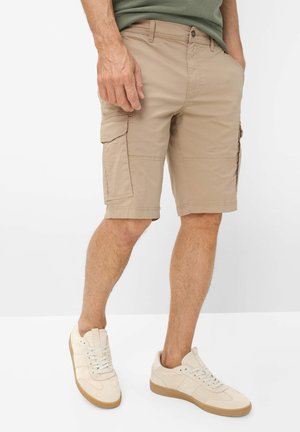 Mann trägt beige Cargo-Shorts und cremeweiße Sneakers, steht mit einer Hand in der Tasche vor weißem Hintergrund.
