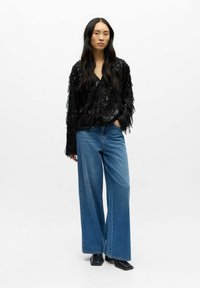 Žena s dlouhými černými vlasy, oblečená v černém flitrovém topu s třásněmi, širokých modrých džínách a černých botách se čtveratou špičkou, stojí na bílém pozadí.