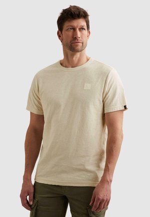 Man die een beige textuur t-shirt met korte mouwen en olijfgroene cargobroek draagt, staand tegen een effen lichte achtergrond.