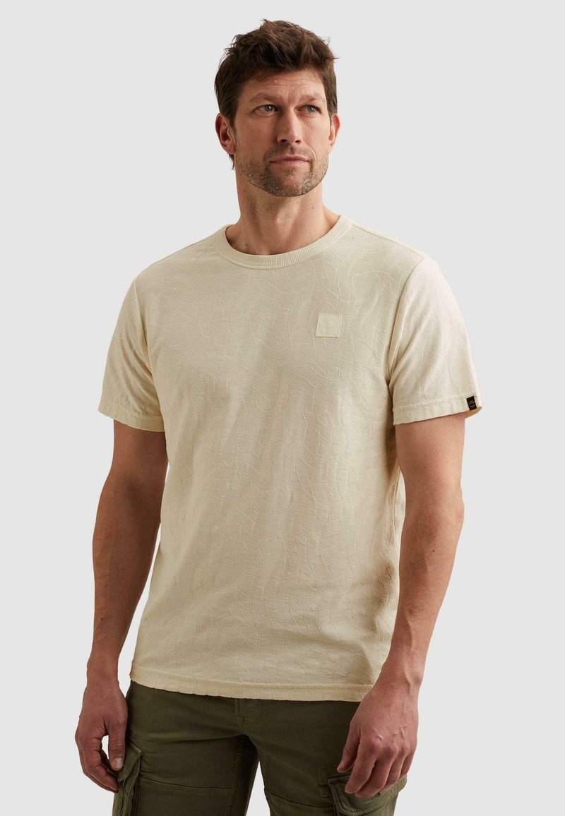 Homme portant un t-shirt beige texturé à manches courtes et un pantalon cargo vert olive, debout devant un fond clair uni.