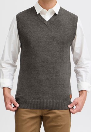 Hombre que lleva un chaleco de punto gris oscuro con cuello en V sobre una camisa blanca abotonada y pantalones marrones, sosteniendo el borde del chaleco con ambas manos.